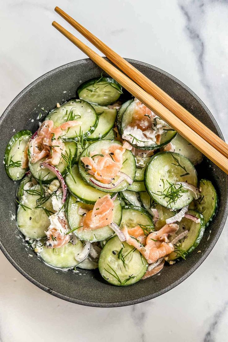 a bowl filled with cucumbers and salmon on top of a marble table next to chopsticks