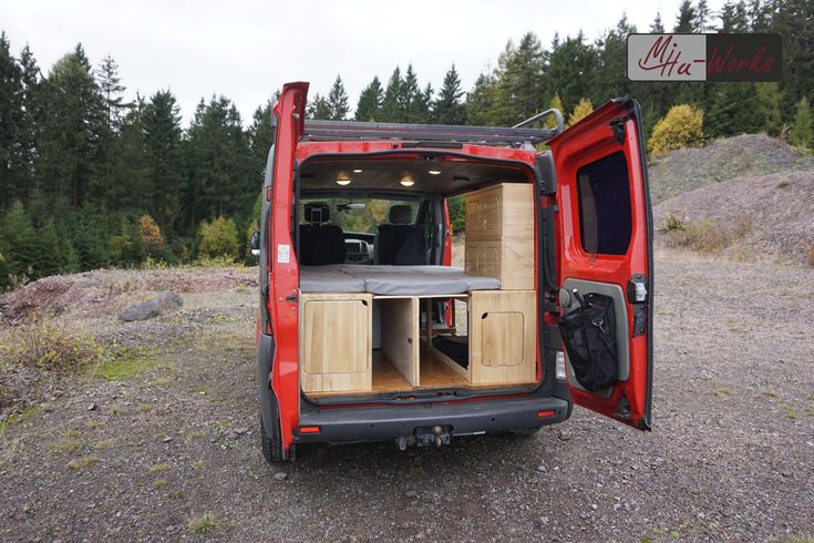 an open red van parked on top of a dirt field next to trees and bushes