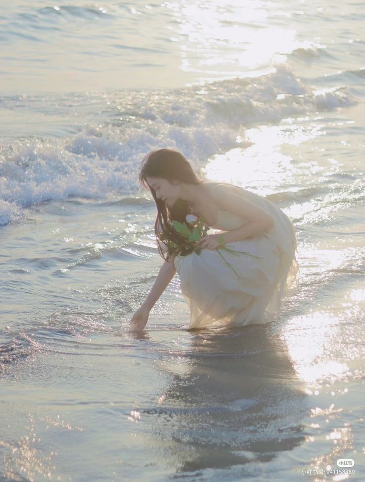 a woman in a white dress is kneeling in the water at the edge of the ocean