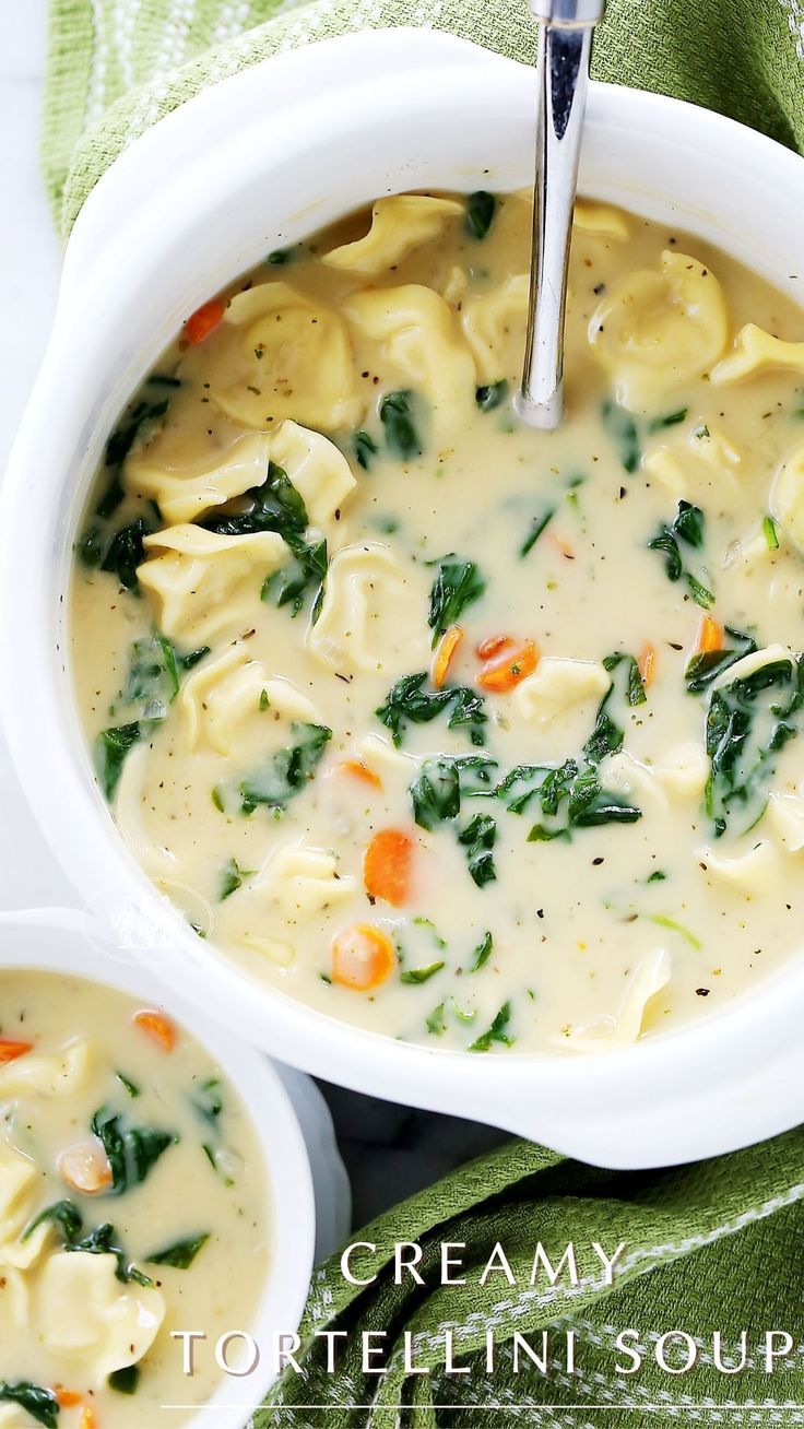 two white bowls filled with tortellini soup on top of a green cloth next to silver spoons