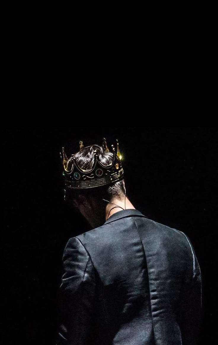 a man with a crown on his head standing in front of a black background and wearing a suit