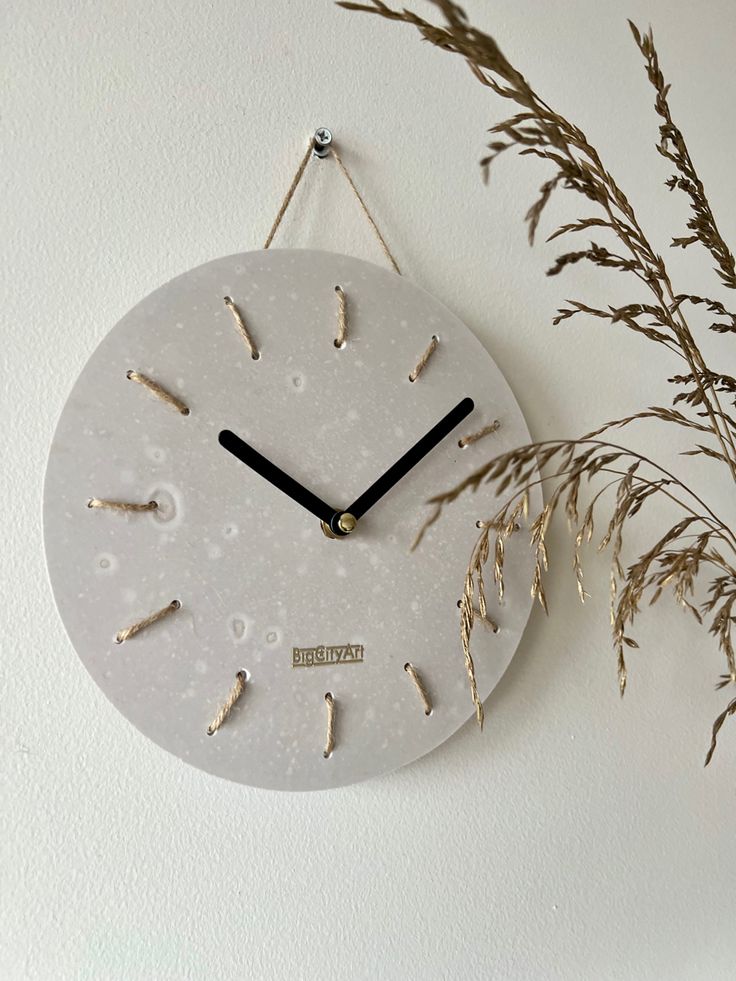 a white clock hanging on the wall next to some dry grass and dried plant stems