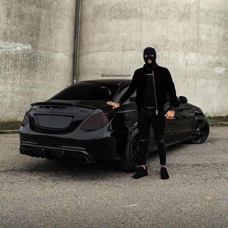 a man standing next to a black car in front of a concrete wall with the hood up