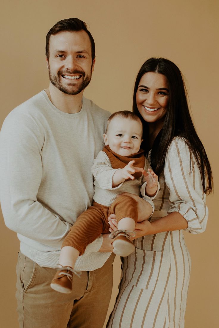 a man and woman holding a baby in front of a beige background with the child smiling