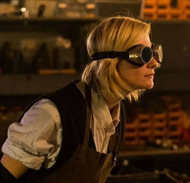 a woman wearing goggles and an apron looks off into the distance in front of shelves