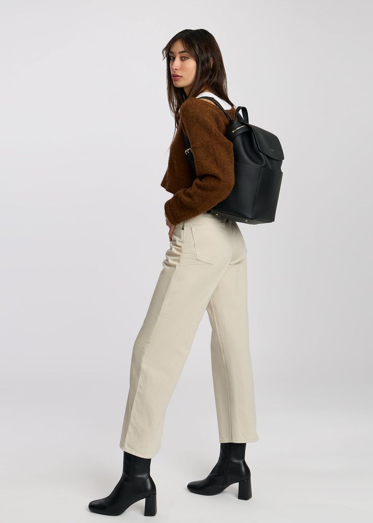 a woman in brown sweater and white pants carrying a black backpack