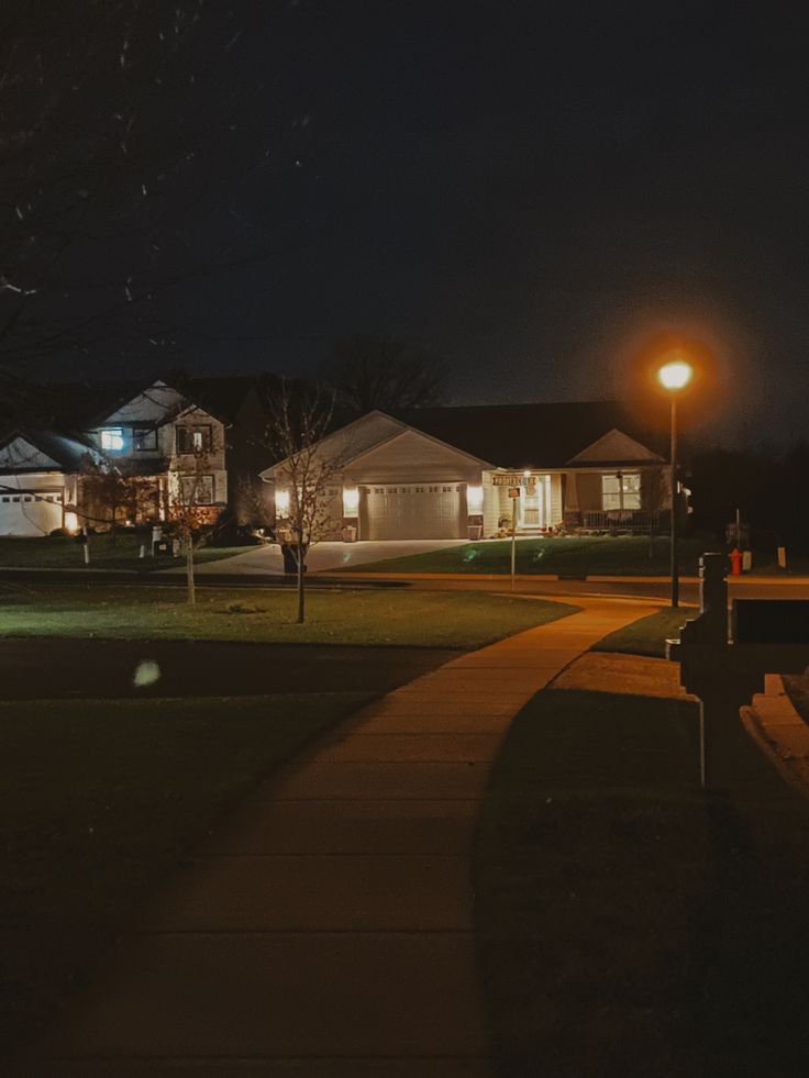 a street at night with houses lit up in the background and lights shining on the grass