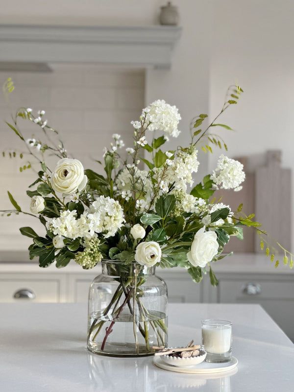 a vase with white flowers and greenery on a kitchen counter next to a candle