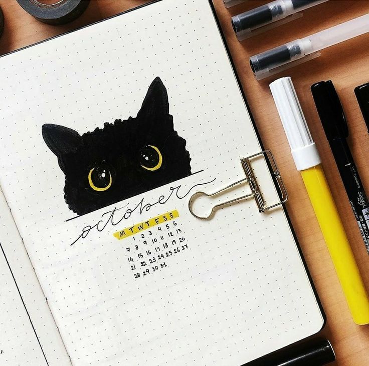 an open notebook with a black cat drawn on the front and yellow eyes, surrounded by other office supplies