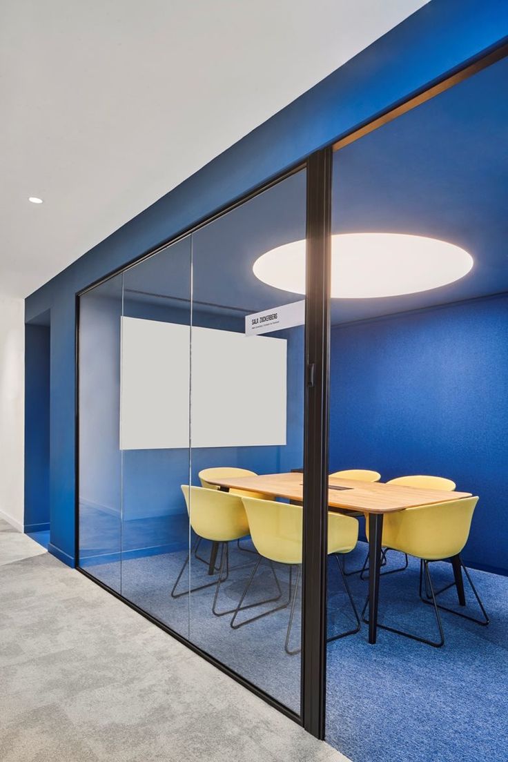 an office meeting room with blue walls and carpeted flooring, yellow chairs are in front of the glass wall