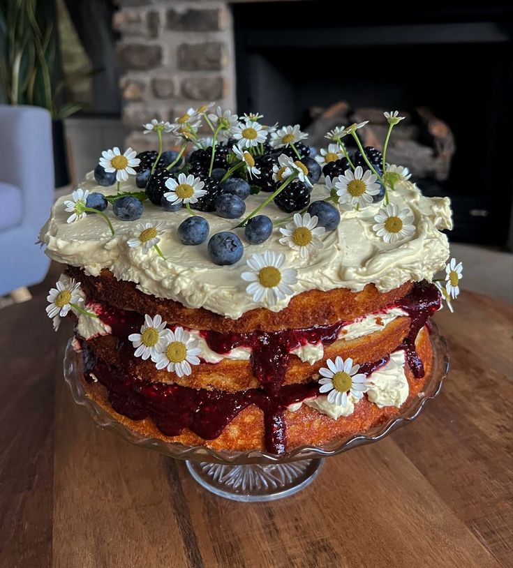 a cake with blueberries, cream and daisies on top sits on a table in front of a fireplace