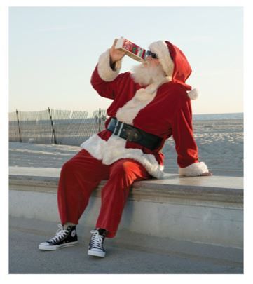 a man dressed as santa claus drinking from a bottle while sitting on a ledge near the ocean