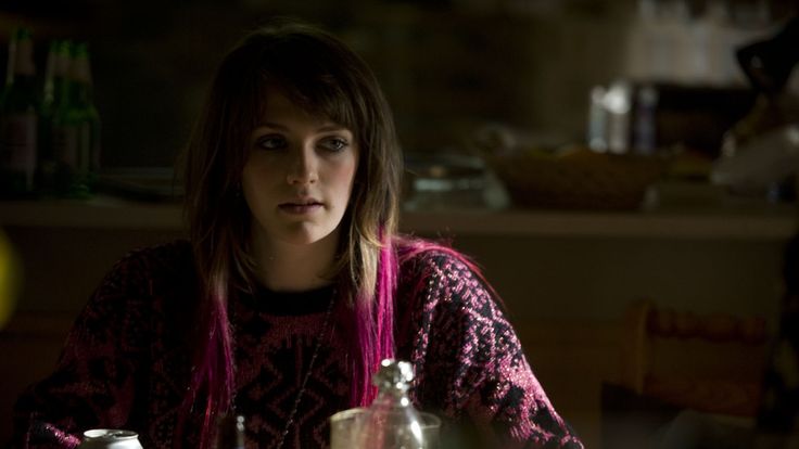 a woman with pink hair sitting at a table in a dark room next to bottles