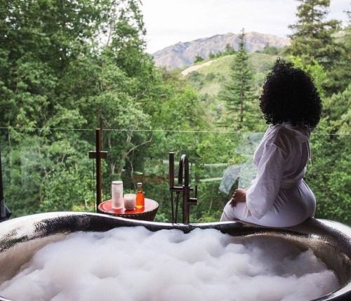 a woman sitting in a bathtub filled with soap next to a forest and mountains