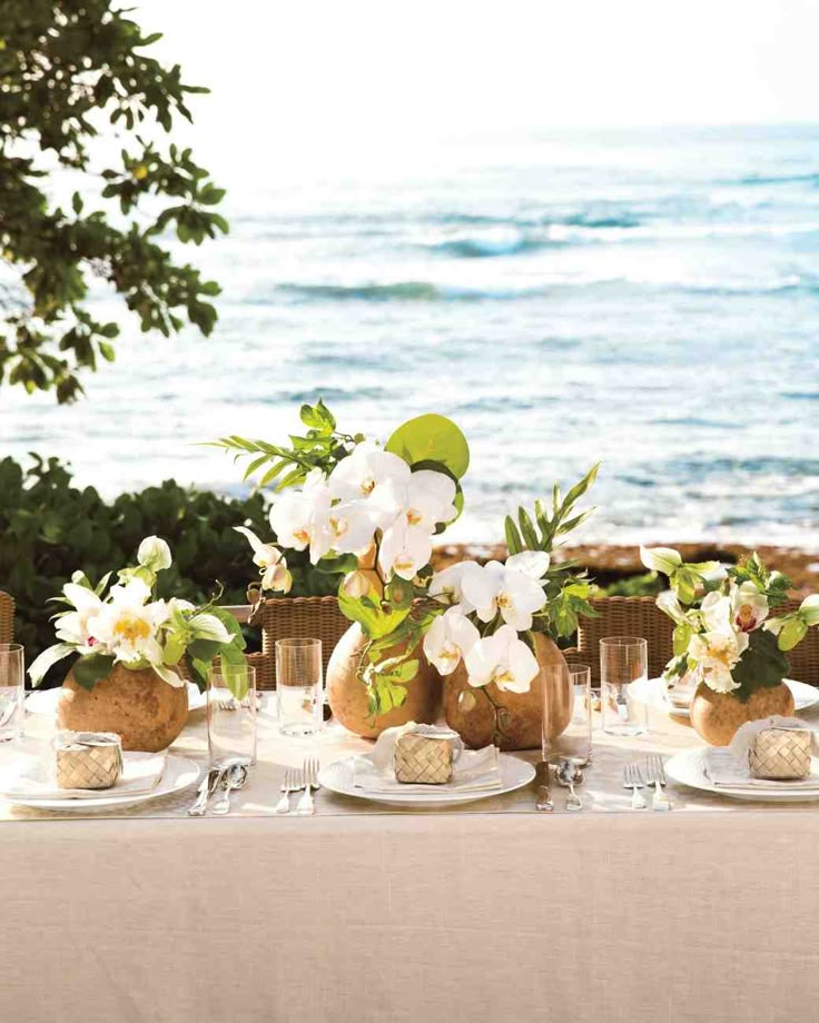 a table set with plates and flowers on it near the ocean in front of some trees