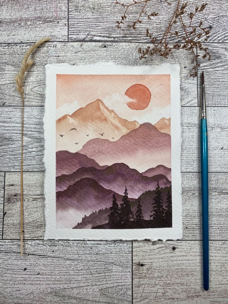 a watercolor painting of mountains and trees on a wooden surface with a pencil next to it