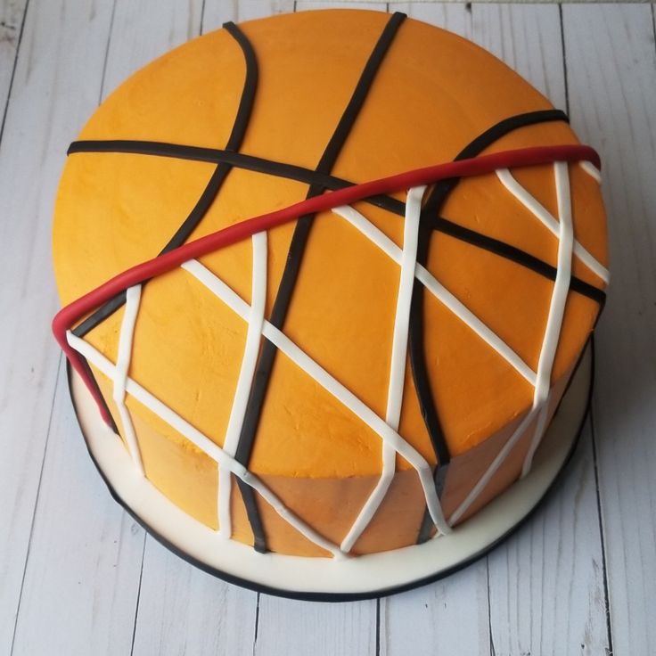 a basketball cake with white and brown icing on a wooden table next to a red ribbon