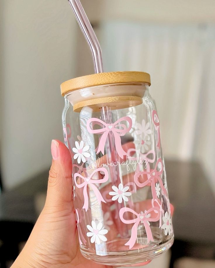 a person holding a mason jar with pink and white flowers on it, while wearing a straw