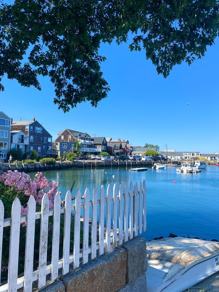 a white picket fence next to a body of water with houses in the background and boats on the water