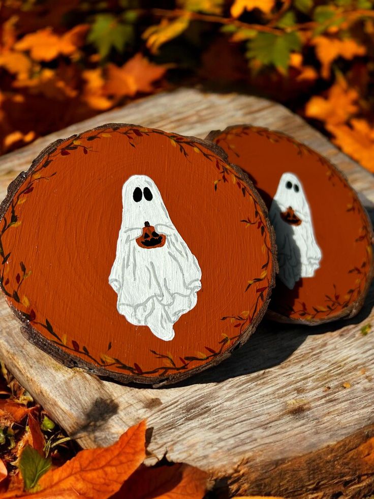 two wooden slices with painted ghost faces on them, sitting on top of fallen leaves