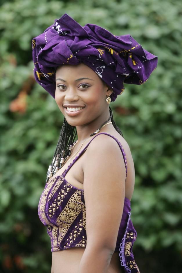 a woman in a purple dress and head wrap smiles at the camera while standing next to some bushes