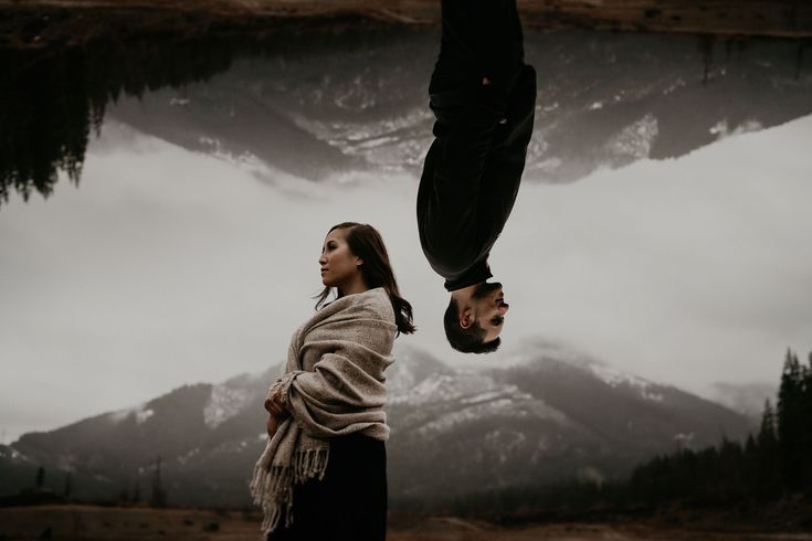 a woman standing next to a man upside down in front of a lake with mountains behind her