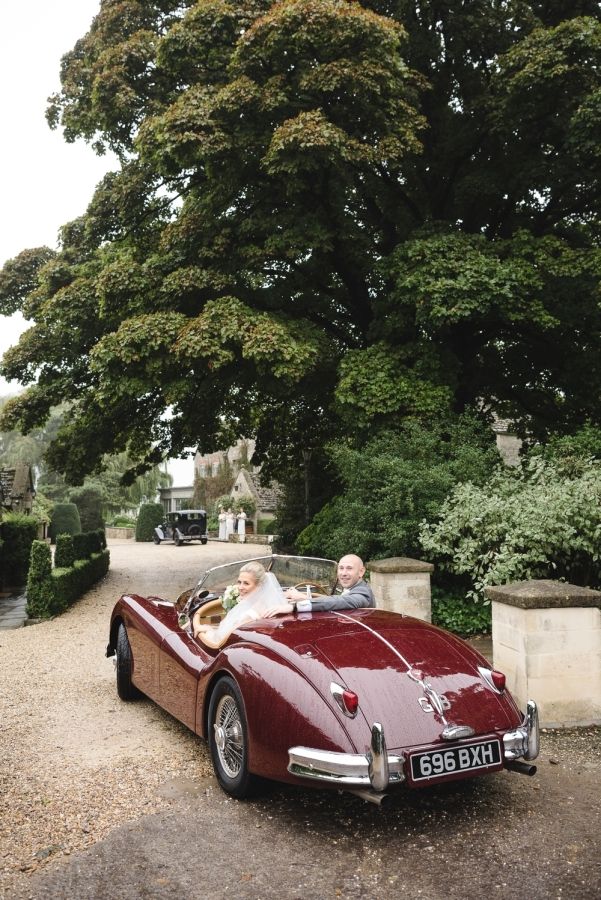 an old man and woman in a red convertible car on gravel road next to trees