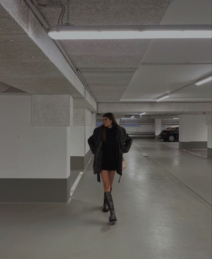 a woman is walking in an empty parking garage with her coat over her shoulders and boots on