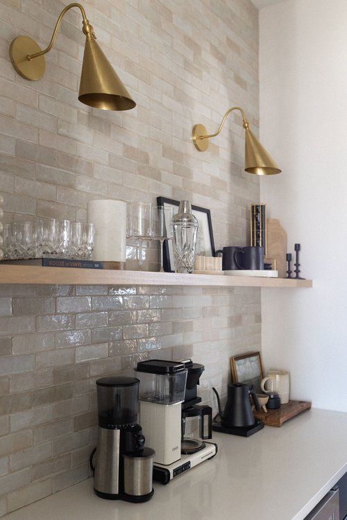 a kitchen counter with coffee maker, cups and glasses on the shelf above it in front of a brick wall