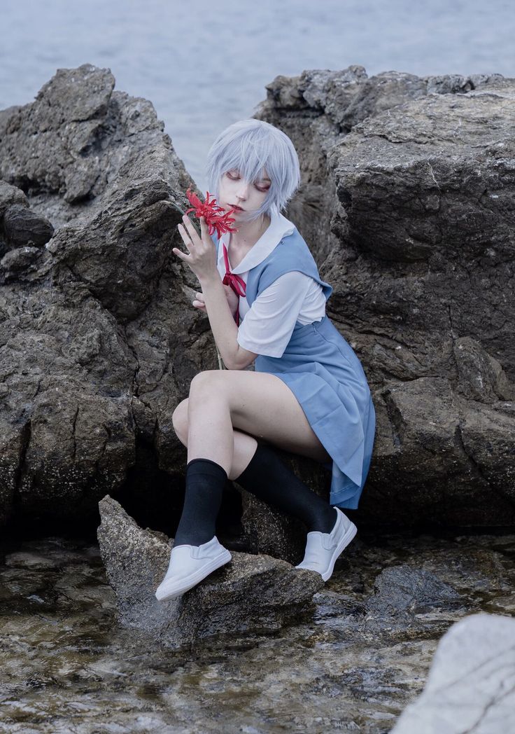 a woman sitting on top of a rock next to the ocean holding a red flower