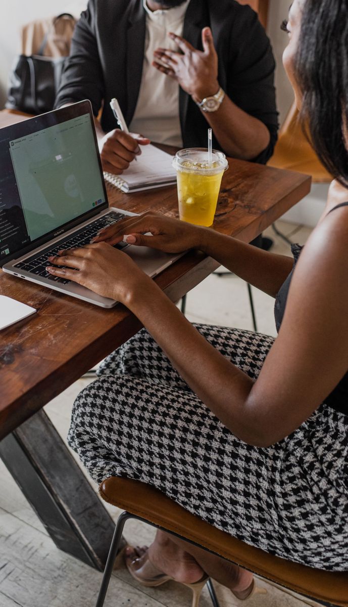 two people sitting at a wooden table with laptops and drinks in front of them