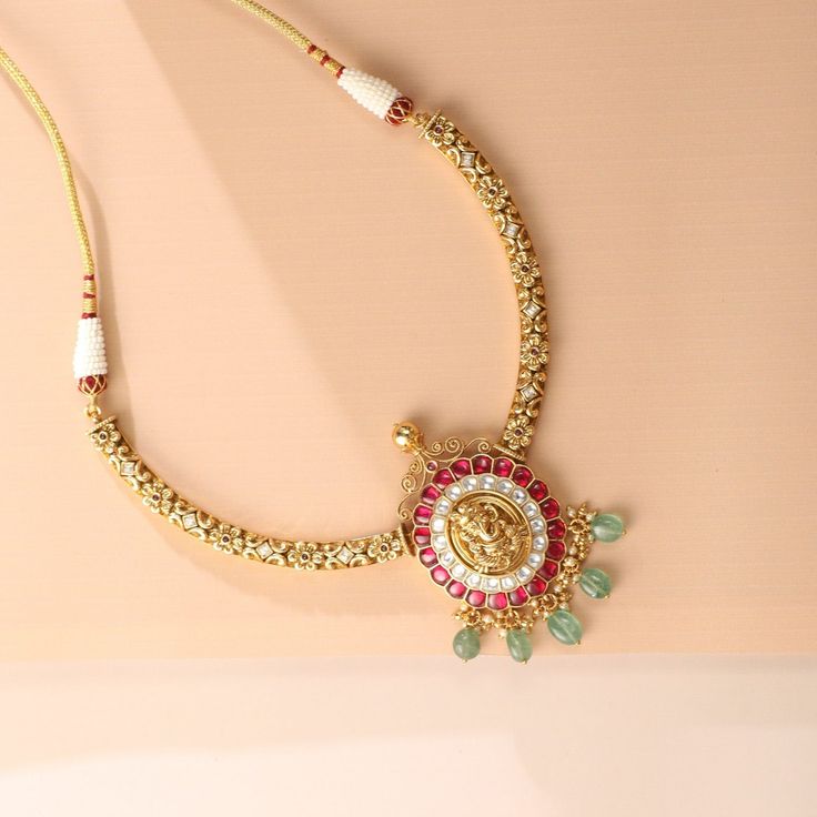 Description: Cascading demurely down the feminine cleft, this elegant hasli combines classic temple patterns of flora and ivy with a handcrafted nakshi depicting Lord Ganesha motif at the apex. Kempu stones set in jadau style and gemstone beads drop adorn the necklace to create a symphony of vivid colors in this unique piece. Product Information Materials used: 925 Silver with 1.0-microns Antique Gold Plating Stones: Semi precious stones Length: 13.5 cm Findings: Hook & links Kundan Temple Necklace For Puja, Elegant Bridal Necklace With Peacock Design For Puja, Elegant Temple Necklace With Zari Work For Navratri, Navratri Necklaces With Zari Work, Festive Temple Jewelry Necklace With Intricate Design, Festive Temple Necklace With Intricate Design, Festive Bridal Necklace With Intricate Design For Puja, Elegant Temple Necklace For Rituals And Festive Occasions, Navratri Temple Jewelry Necklace With Zari Work