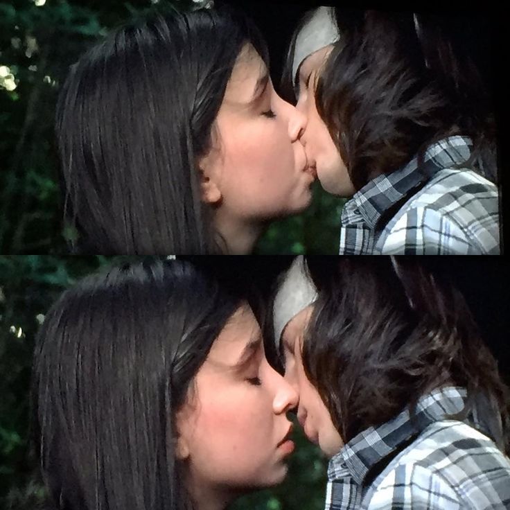 two women are kissing each other in front of some trees and bushes, one is wearing a plaid shirt