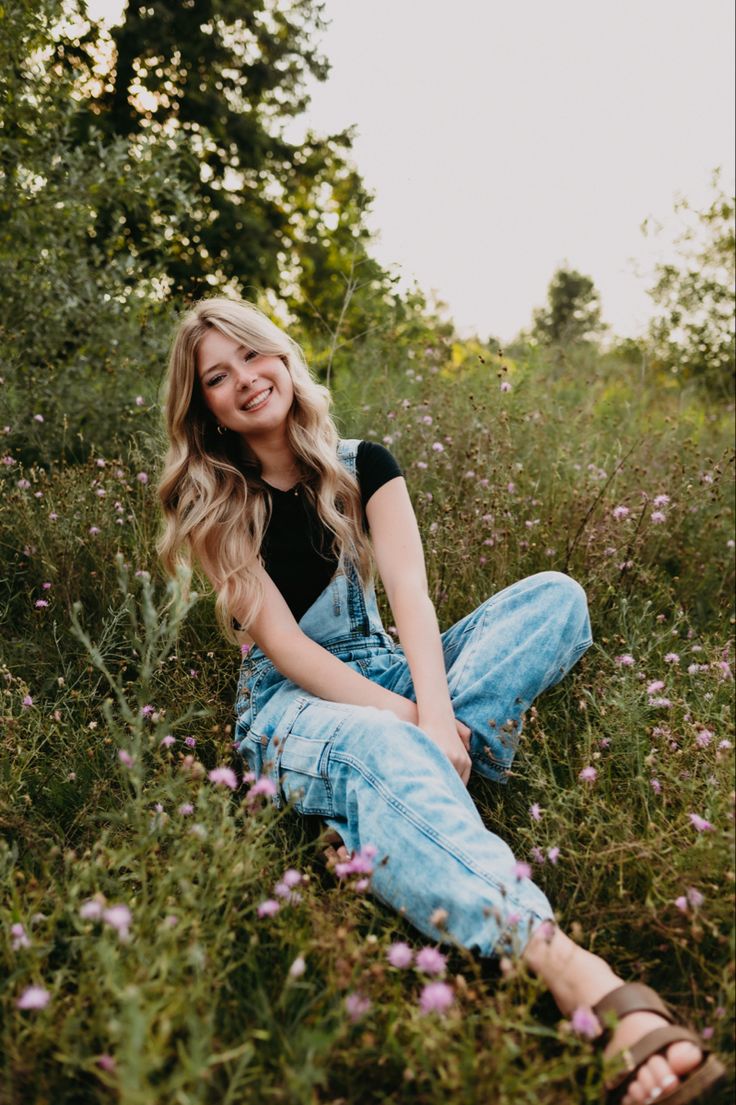 a woman sitting in the middle of a field with wildflowers and trees behind her