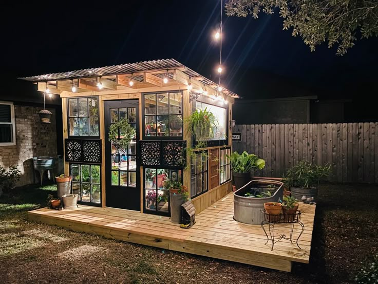 a small garden shed lit up at night with lights on the roof and potted plants outside