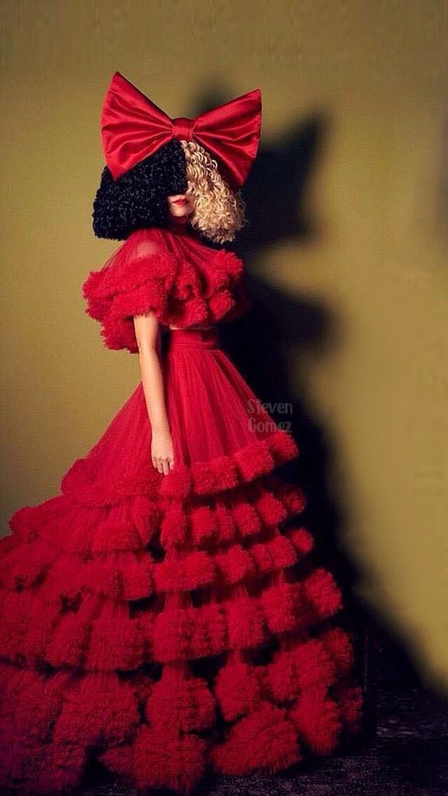 a woman in a red dress with a big bow on her head, standing next to a wall