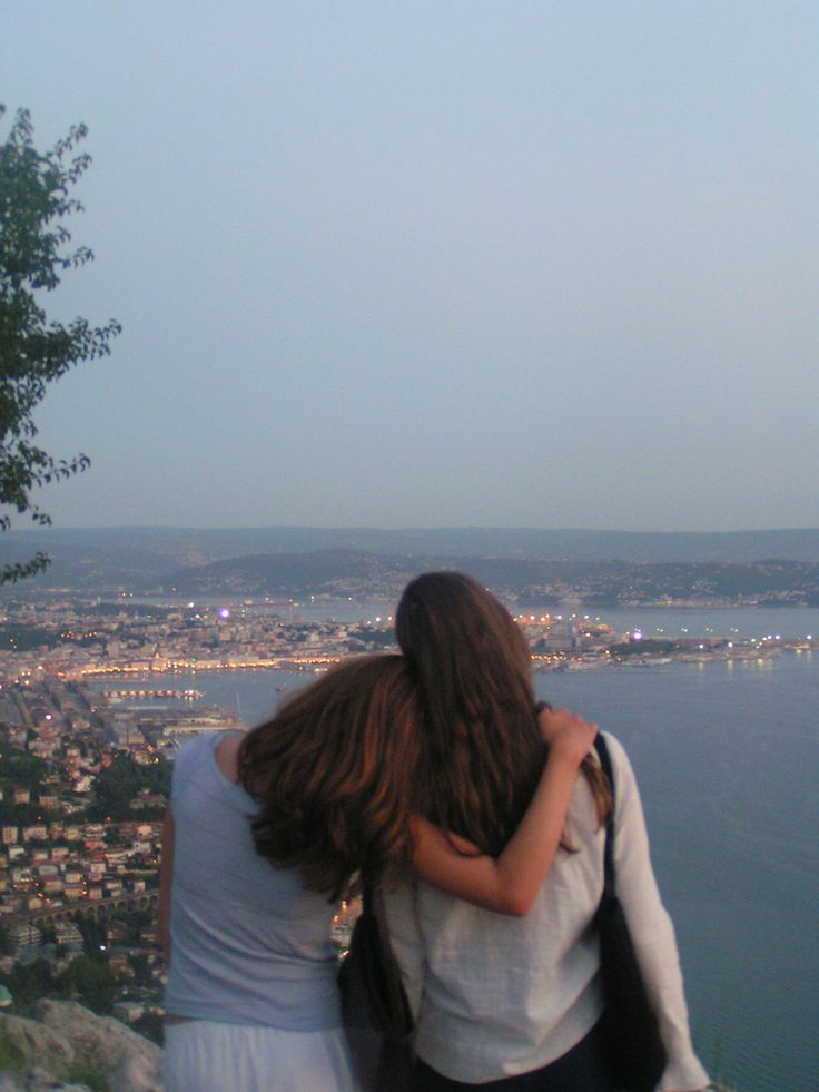 two people standing on top of a hill looking at the water and city below them