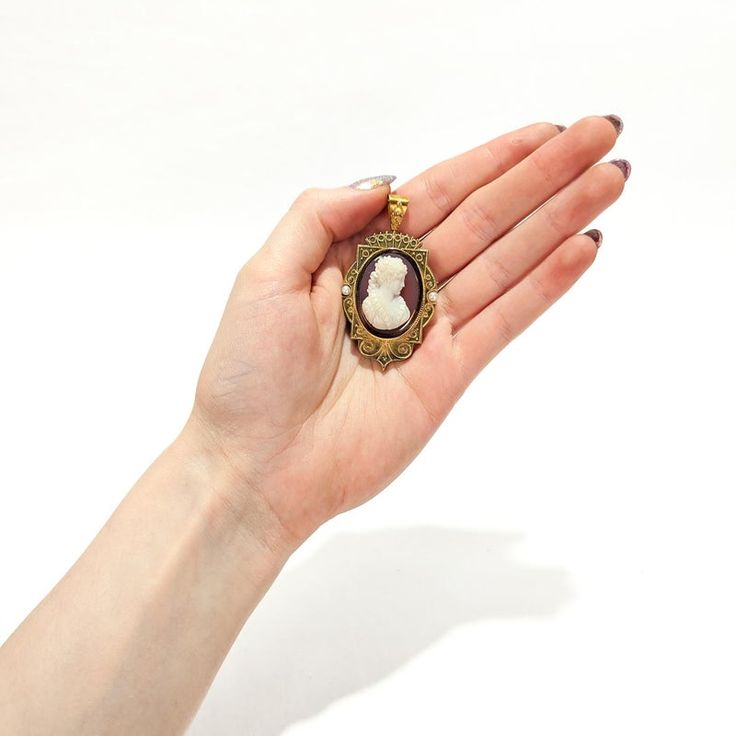 A fine antique Victorian convertible brooch / pendant for a necklace.  In 14k gold.  Comprising a cameo brooch with a oval reserve for a photo to the reverse and a tapered bail.   Centered on a pink and white carved agate or hard stone cameo of a beautiful young woman with flowing curly hair, earrings, and a ruffled dress.   The sides are set with small, round pearls and there is applied twisted rope and granulated decoration throughout.  Simply a wonderful piece of Victorian jewelry!  (Chain for display purposes only and not included with purchase).  Date: Mid-19th Century  Overall Condition: It is in overall good, as-pictured, used estate condition. There is some light edge wear, some fine and light surface scratches, and other signs of light wear consistent with age.  Fineness: Unmarked Oval Cameo Jewelry For Memorial, Oval Cameo Brooches As Gift, Victorian Pendant Brooches With Cabochon, Victorian Brooch Jewelry For Memorial, Victorian Brooch For Memorial, Victorian Cameo Jewelry For Memorial, Victorian Brooch For Memorials, Victorian Cameo Jewelry For Memorials, Oval Cameo Jewelry Gift