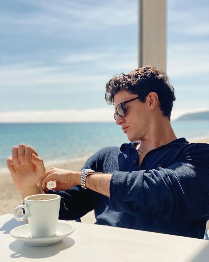 a man sitting at a table with a cup of coffee in front of him on the beach