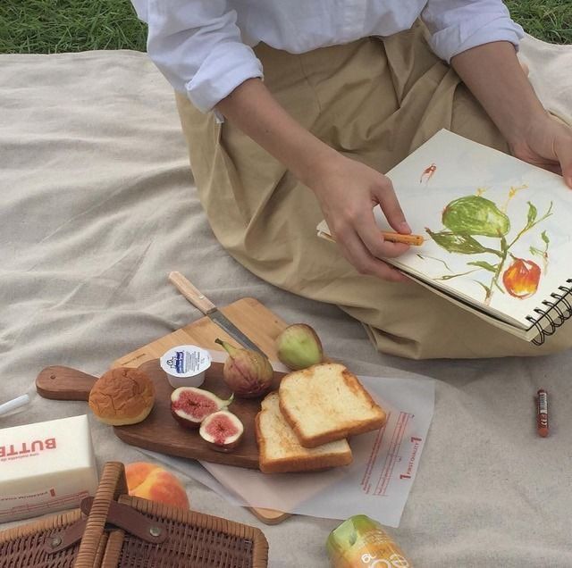 a woman sitting on the ground with bread, fruit and an open book in her hands