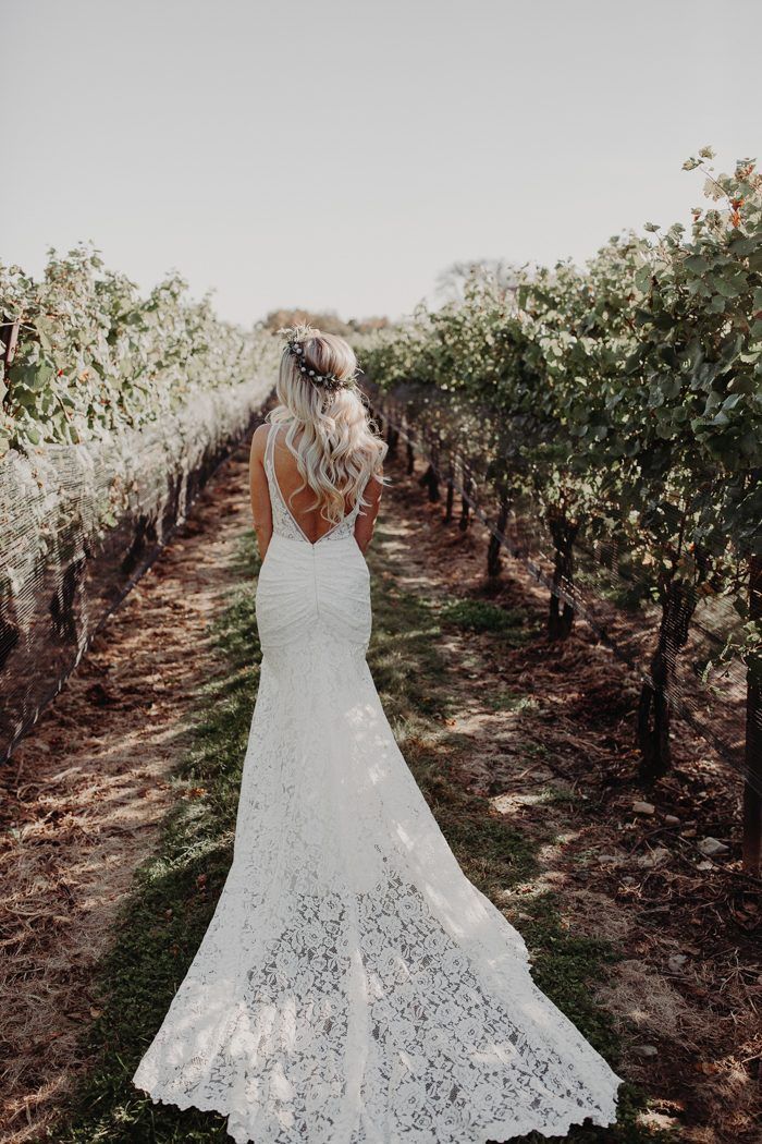 a woman standing in an orchard wearing a white lace wedding dress and holding her hair back to the camera