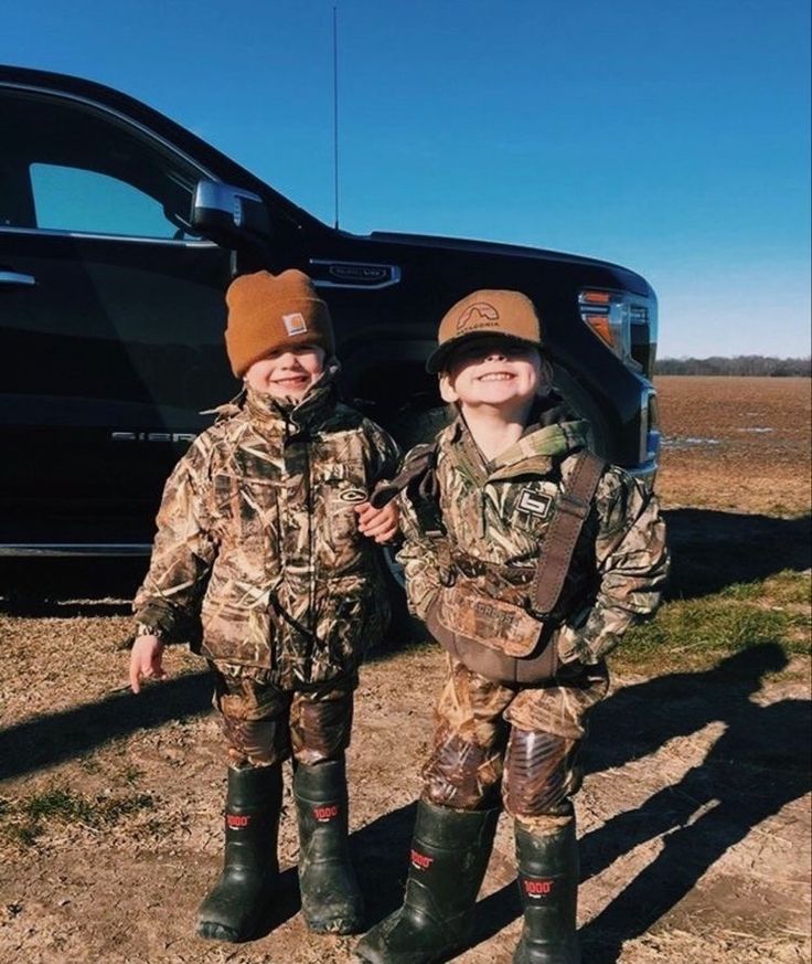 two young boys standing next to each other in front of a black truck with camouflage gear on