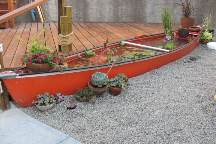 an orange boat filled with plants on top of a gravel ground next to a wooden deck