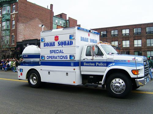 a white and blue truck driving down a street next to tall buildings with people in the background