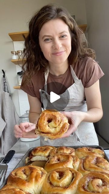 a woman holding up a pastry in front of a pan filled with cinnamon buns