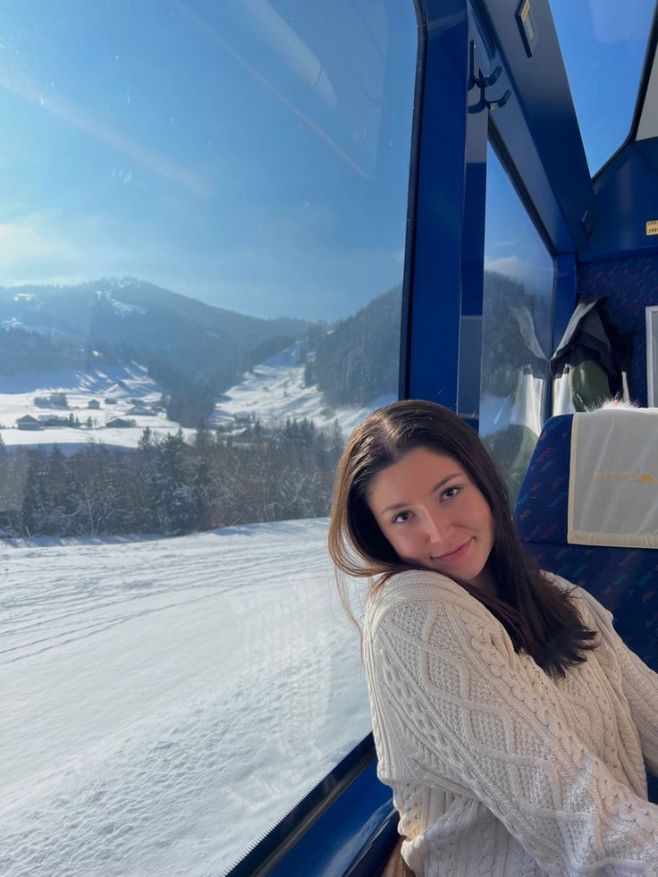 a woman sitting on a train looking out the window at snow covered mountains in the distance