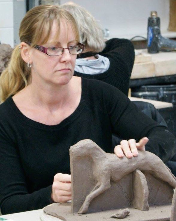 a woman sitting at a table working on a clay horse sculpture with another woman in the background