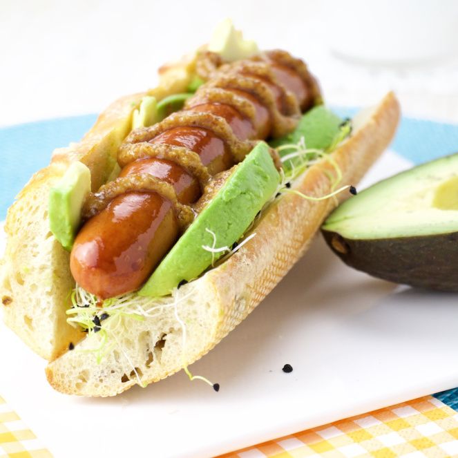 a hot dog on a bun with avocado and other toppings next to an avocado