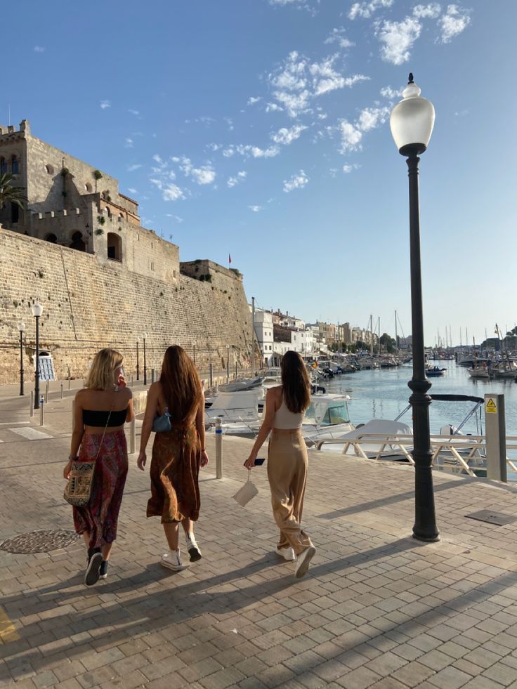 three women walking down the street in front of an old city wall with boats on it