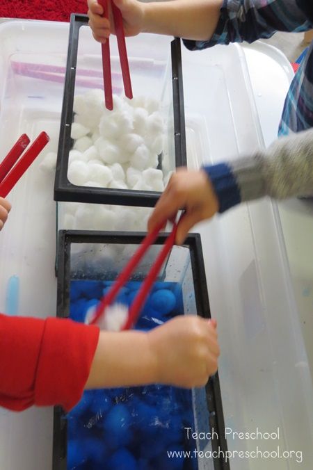 two children are playing with red chopsticks in a plastic container filled with white cotton balls